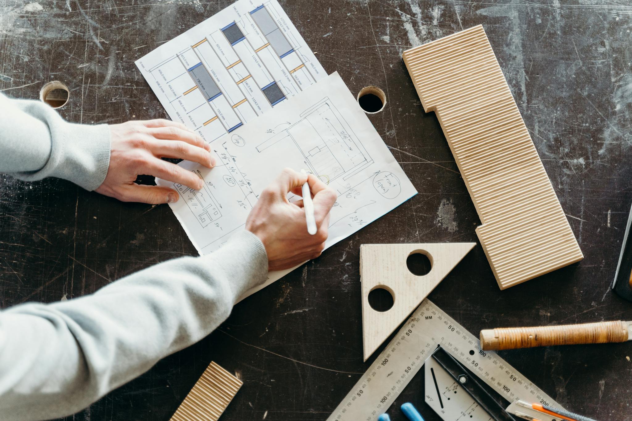 Hands drafting architectural designs on paper with wooden tools in a workshop setting.
